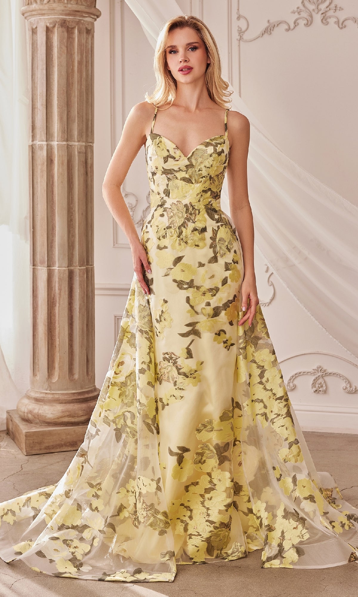 A woman poses indoors in the Lemon Yellow Floral-Print Long Prom Dress A0770, featuring a fitted bodice and flared train. The elegant floral formal dress flows gracefully as she stands near a column with draped white fabric.